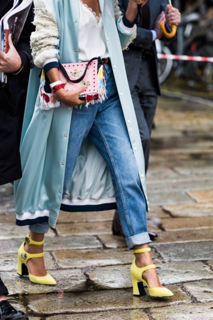 close up of woman's torso wearing rolled up jeans, yellow heels with wide straps, white lacy top and light blue over-sized bomber trench coat with fluffy sleeves, holding studded and tasseled clutch 