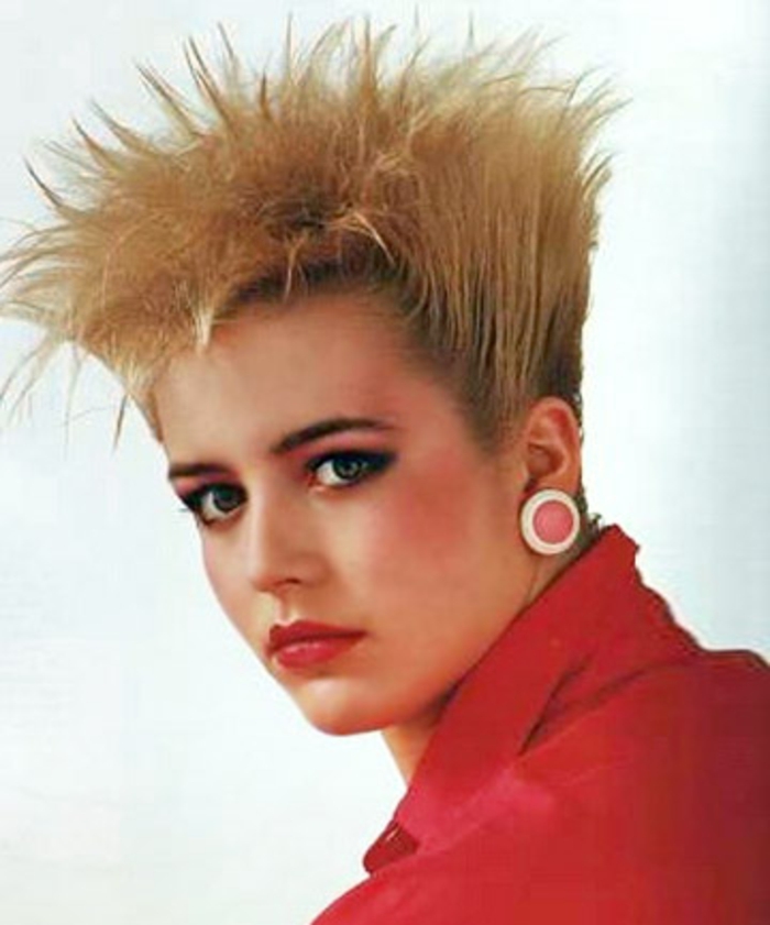 close up of blonde woman with 80s gelled up hi-top fade hair, red shirt and big round clip-on earring, red lipstick and heavy make up