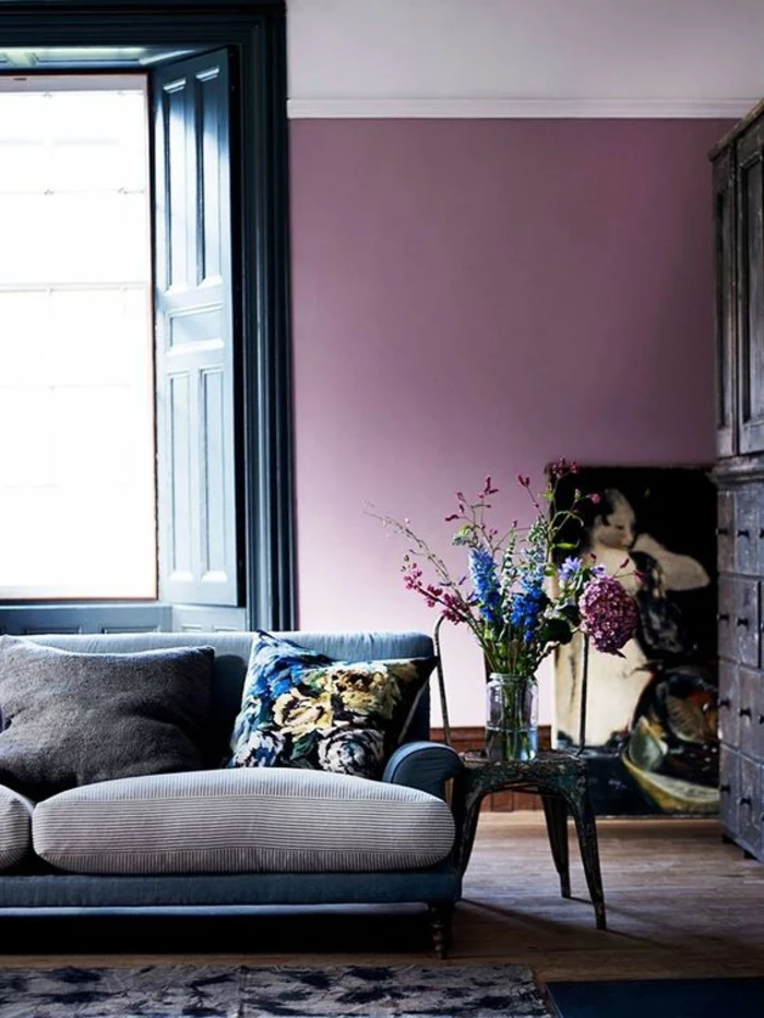interior painting, purple wall, grey and black sofa with colorful cushion, black open door and brown cupboard, dark wooden chair, small dark table with clear jar containing flowers
