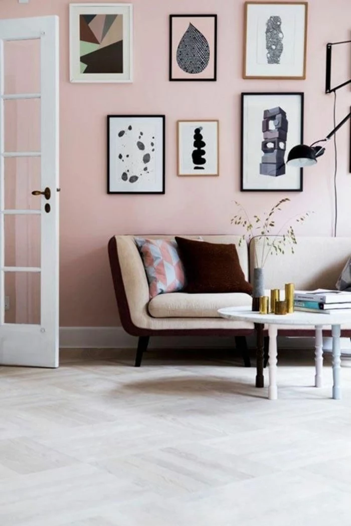 pink wall with six framed images, pale cream and brown sofa with two cushions, pale grey floor and open white door with glass inserts, small table with pastel colored legs