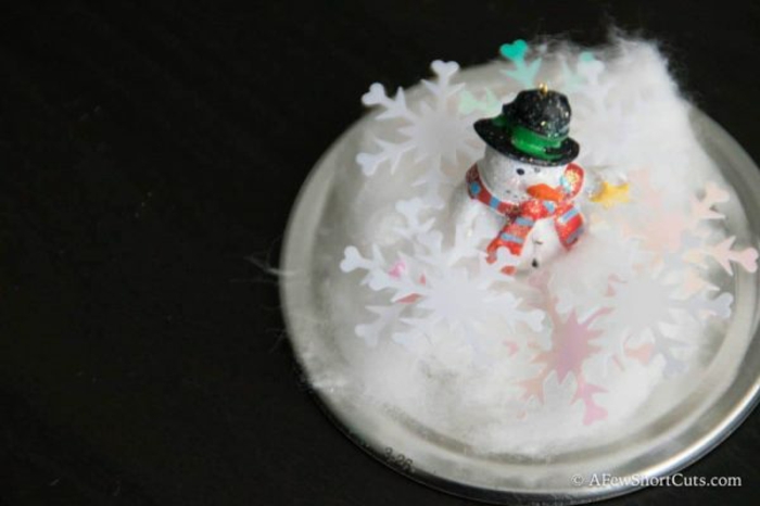 diy christmas gifts, jar lid with cotton, white and pink snow-shaped confetti, a small snowman figurine with hat and scarf, black background