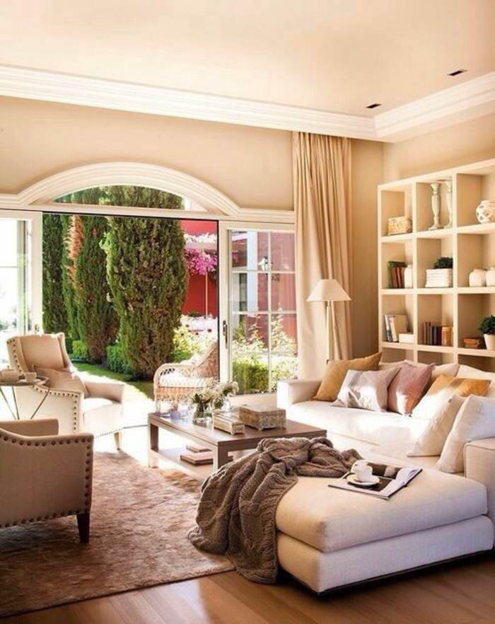 living room with pale orange walls and ceiling, white plaster details, cream sofa and chairs, light brown carpet and wooden floor, white shelves and cream curtains, grey blanket and glass door opened to garden