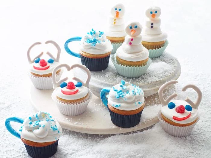 batch of cupcakes with white frosting, in white dark blue and light blue wrappers, decorated with candies to look like snowmen and cups
