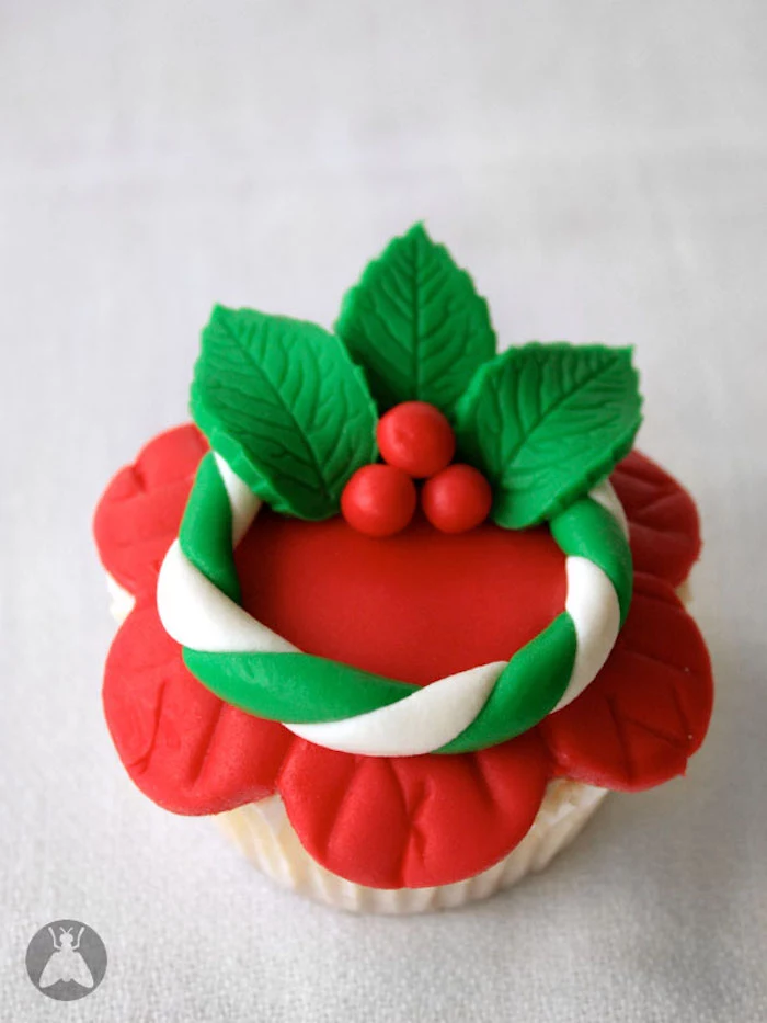 cupcake with red fondant icing shaped like flower, decorated with white and green fondant wreath with holly