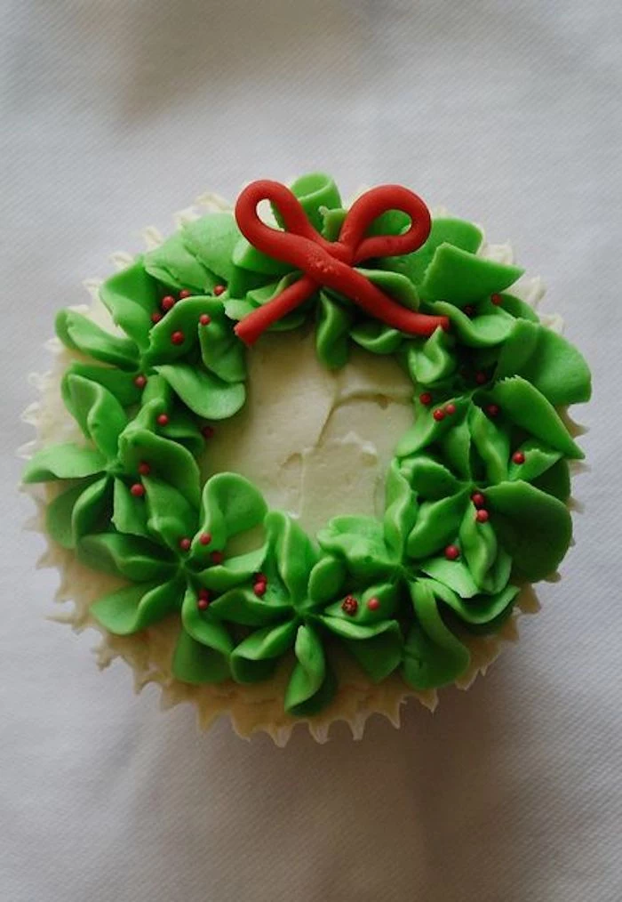 close up of cupcake with white icing, made to look like an x-mas wreath with green frosting, with small fondant bow and red sprinkles