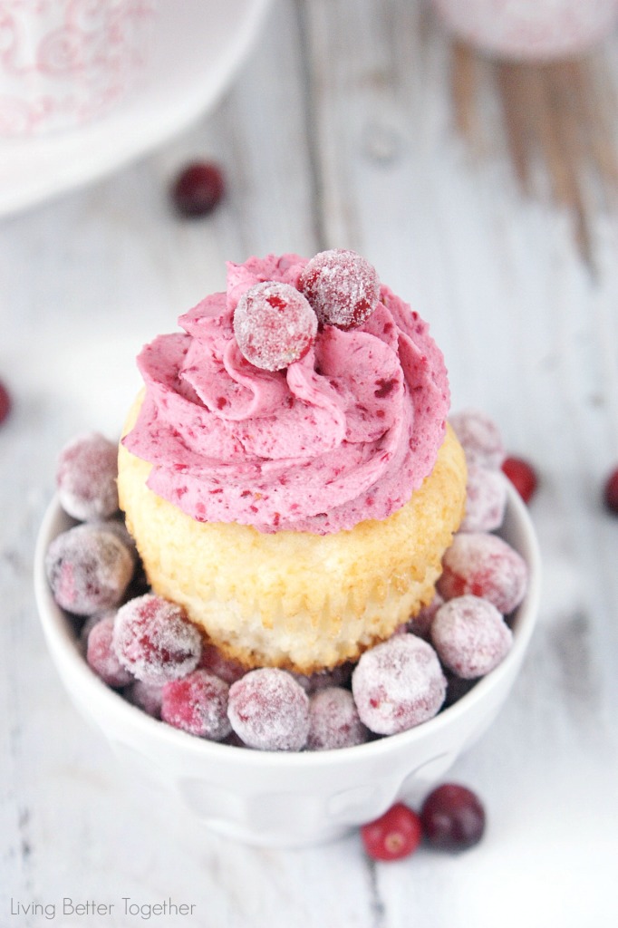 small white dish with frosted cranberries, and a yellow cupcake with pink icing, decorated with frosted cranberries, on white wooden surface