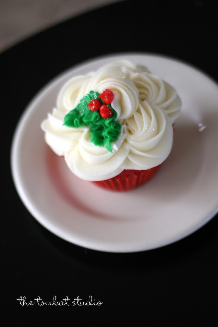 red cupcake in red wrapper, with white frosting, decorated with fondant holly, white plate on black background