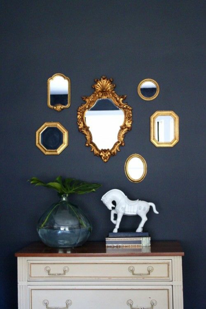 six differently sized and shaped ornate mirrors with golden frames, white chest of drawers with brown top, blue glass vase with green plant, books and a white horse statuette, dark blue background