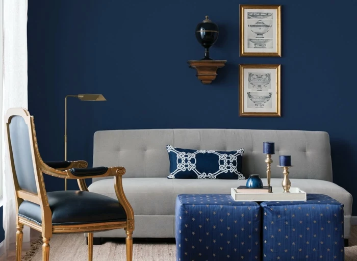 living room color ideas, dark blue wall and light grey sofa, navy blue chair with gold details, two blue ottomans with yellow stars, gold lamp and two framed pictures