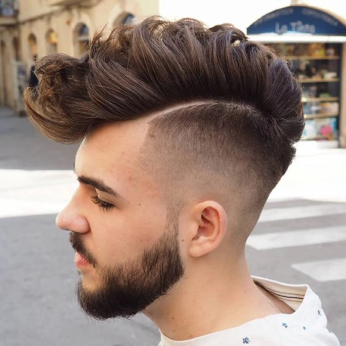 side part undercut, young bearded and mustached man in profile, white top with blue details, dark hair shaven on the side and long on top, gelled up and messy