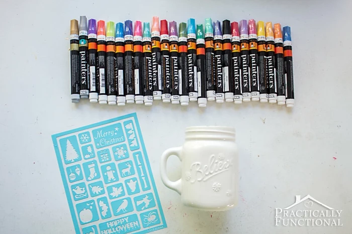 diy christmas crafts, many markers in different colors, near a white empty mug with handle, a blue holiday-themed stencil, on white surface