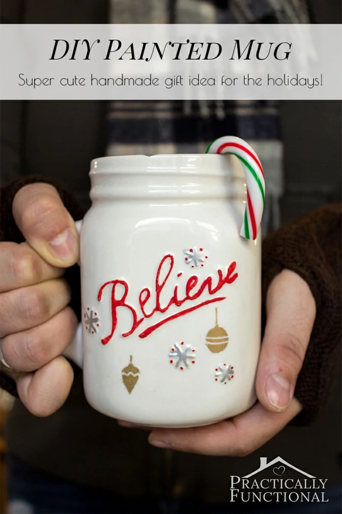 diy christmas crafts, two hands holding a big white mug with handle, with the word believe written in red, hand-drawn snowflakes and Christmas ornaments in blue and gold, a peppermint cane inside
