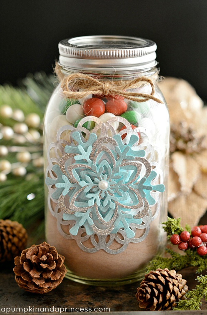christmas crafts for adults, a big clear mason jar filled with sweets, with beautiful snowflake-shaped paper decoration and a pearl, tied with plain string, near pine cones and berries