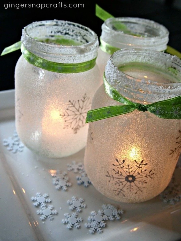 xmas gifts, three frosted mason jars tied with green ribbons, with snowflake drawings and lit candles inside, placed on white tray with snowflake-shaped confetti 