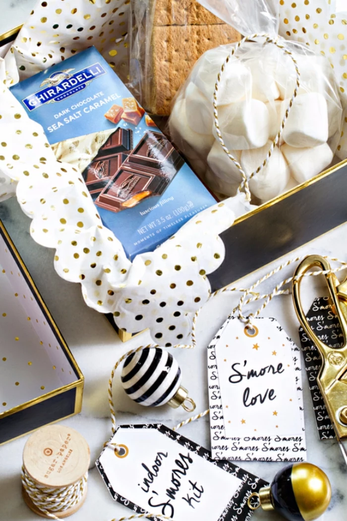 a black and gold box with white and gold wrapping paper inside, with chocolate and biscuits and a bag of white marshmallows, near festive labels and scissors and small christmas ornaments