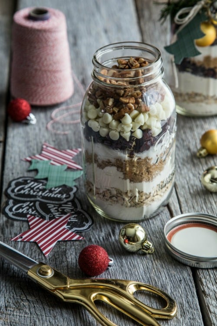 family christmas gifts, open mason jar with walnuts, white chocolate chips and sugar, dried fruit flour and oatmeal, on grey wooden surface near scissors, string, labels and small ornaments