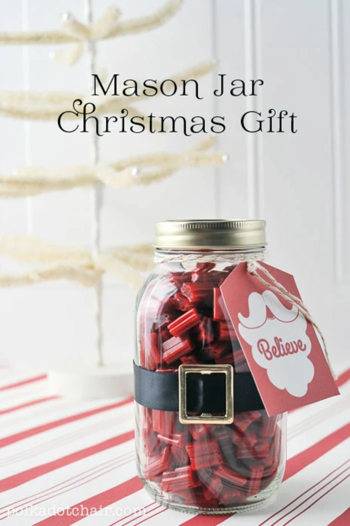 family christmas gifts, a mason jar filled with red candy, a black belt with yellow buckle, and a red festive card, white and red surface with fake white christmas tree in the background