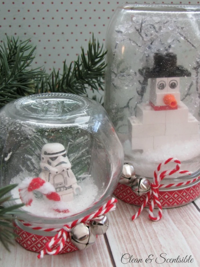 two snow globes made from a small and a large jar, with fake snow and lego figurines, a snowman made of lego, a lego star wars storm trooper, tied with red and white string with two bells each, pine branches in background