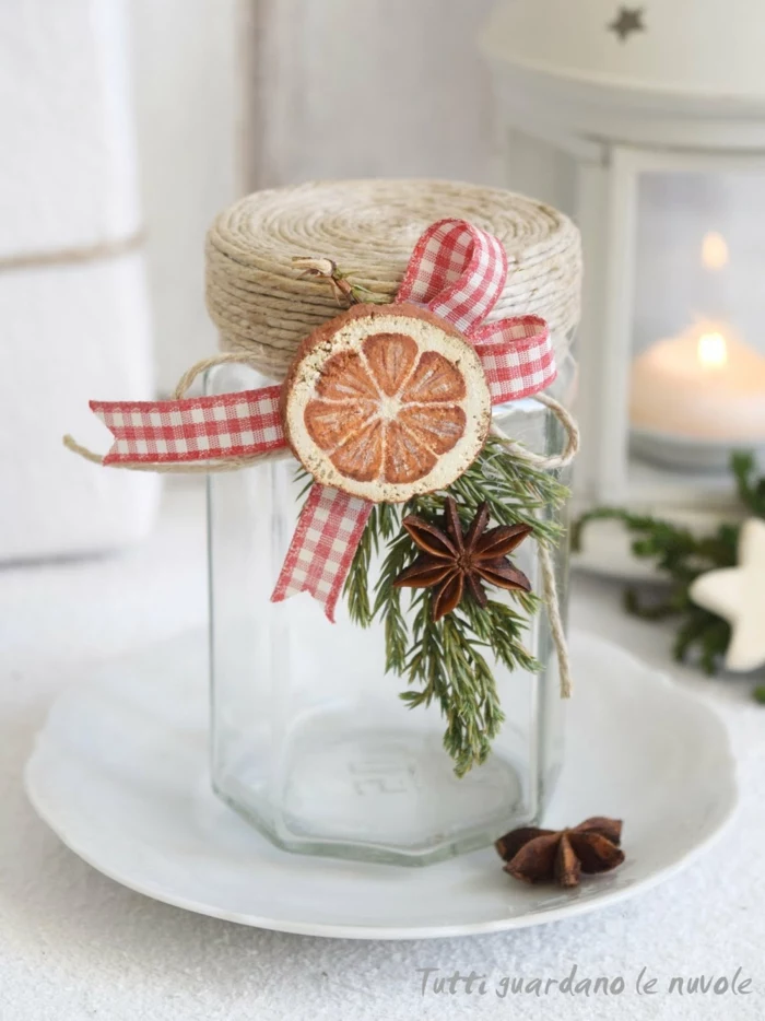 christmas crafts for adults, an empty clear jar decorated with string, red and white checkered ribbon, a wooden ornament painted like an orange slice, star anise and a green twig, placed on a white plate