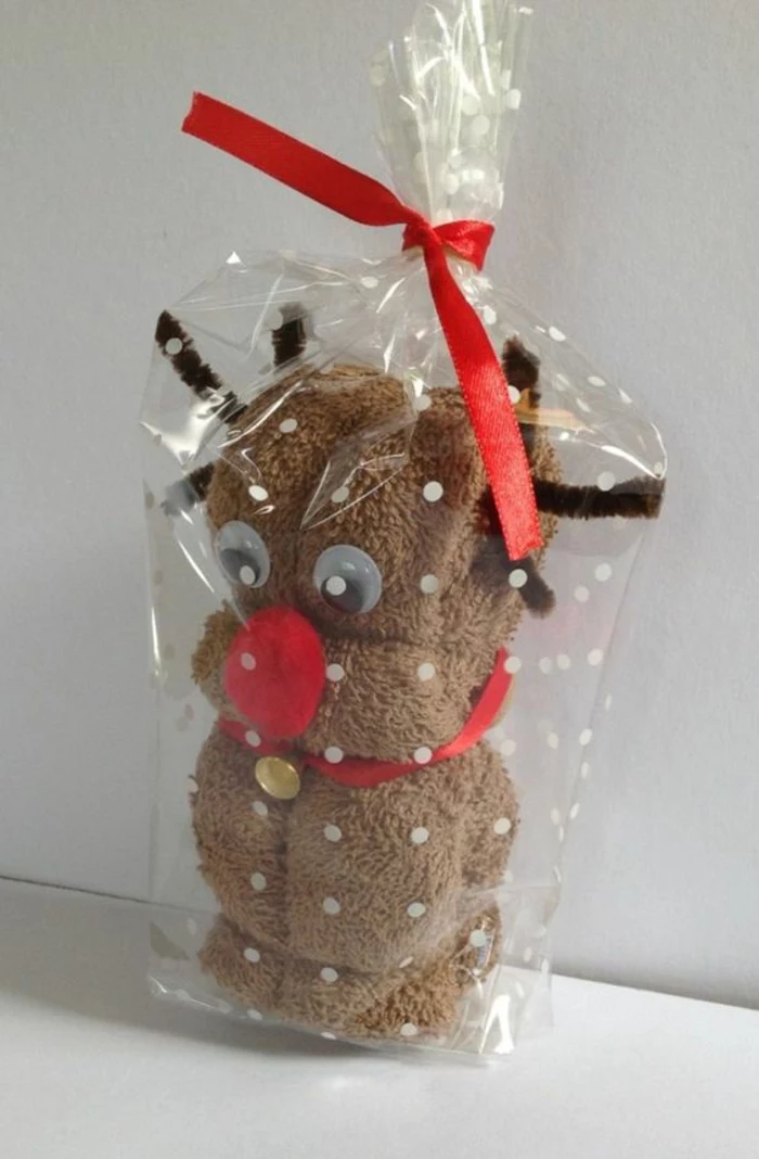 brown fluffy towel made to look like a reindeer, with red collar and a golden bell, googly eyes, brown wire antlers and red pom-pom nose stuck on it, in clear plastic wrapping paper tied with red ribbon