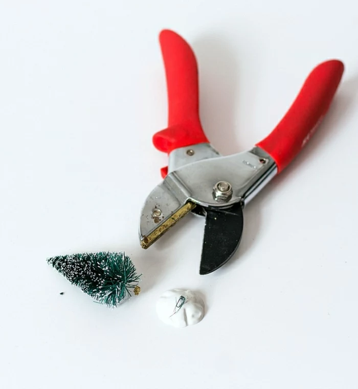 diy gift ideas, pair of metal pliers with red plastic handles, a small snowy Xmas tree ornament, a white plastic stand, white background