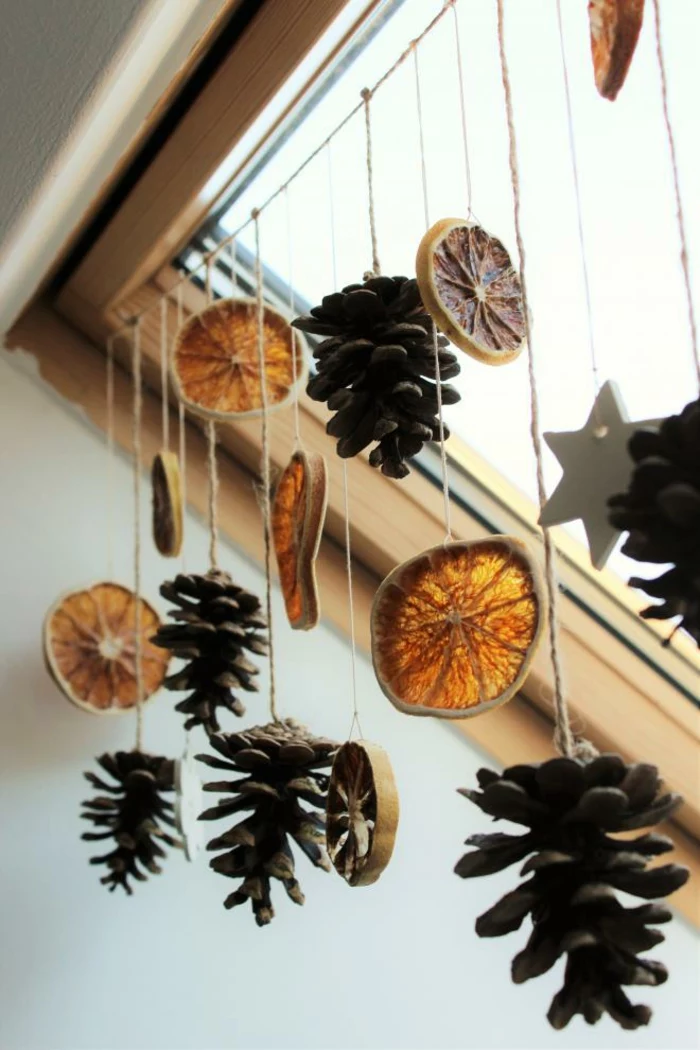 dried orange slices, several pine cones and star shapes, tied to a string and hanging from a ceiling window with wooden window pane
