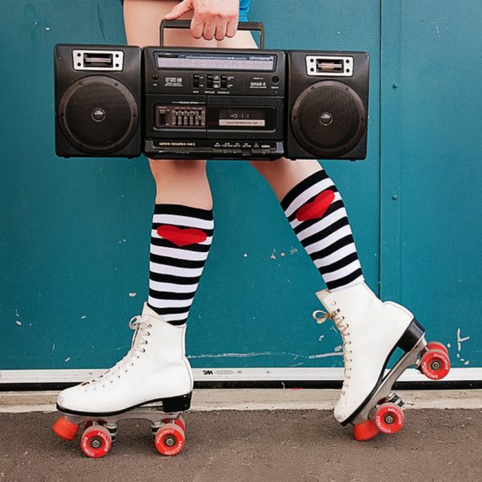 close up of legs wearing retro white skates with red wheels, black and white stripped bellow the knee socks with red hearts, hand holding black retro stereo or boombox, blue background and pavement