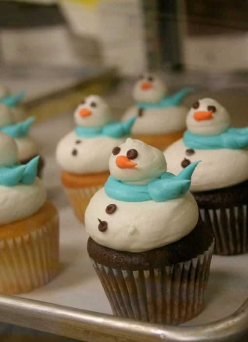 batch of vanilla and chocolate cupcakes, decorated with white marshmallow frosting, shaped like snowman, with pale blue scarf, orange nose and chocolate details