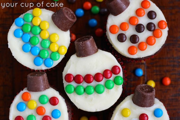 five cupcakes with buttery smooth icing, decorated with chocolates and colorful candies, made to look like christmas tree ornaments