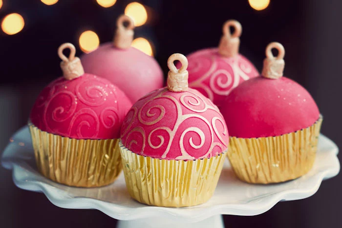 five cupcakes in golden wrappers, with pink fondant icing made to look like christmas tree ornaments, white dish on dark background