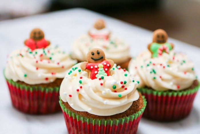 four cupcakes in red and green wrappers, with creamy white icing, decorated with small gingerbread men, and red and green sprinkles