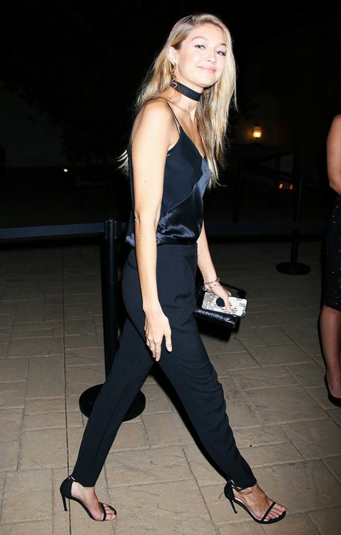 gigi hadid wearing smart black trousers, shiny black strappy top, high heeled sandals and a black satin chocker, holding small black clutch and phone