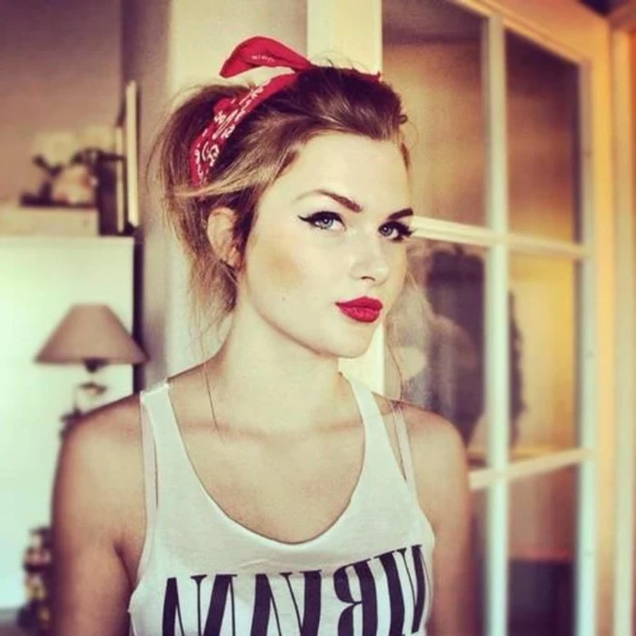 young woman with pinup make up, red lipstick fake lashes and eyeliner, hair tied back with red and white bandanna, wearing white tank top with black writing