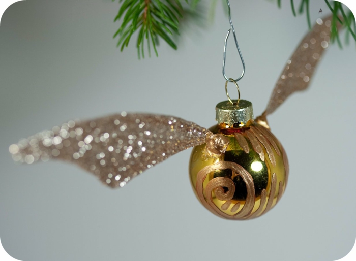 a yellow tree ornament painted with gold paint, with glittering wings and a golden cap, hanging from christmas tree on a metal hook