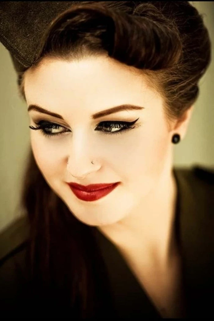 dark-haired smiling woman, deep red lipstick and fake lashes, mascara eyeliner and penciled eyebrows, black earring and black top, nose ring and vintage hairstyle