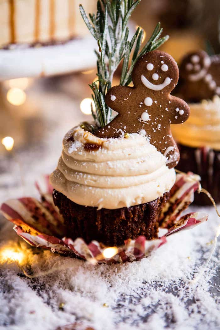 christmas cupcakes, half unwrapped dark cupcake with light brown frosting, decorated with gingerbread man and a rosemary stalk, fake snow and fairy lights