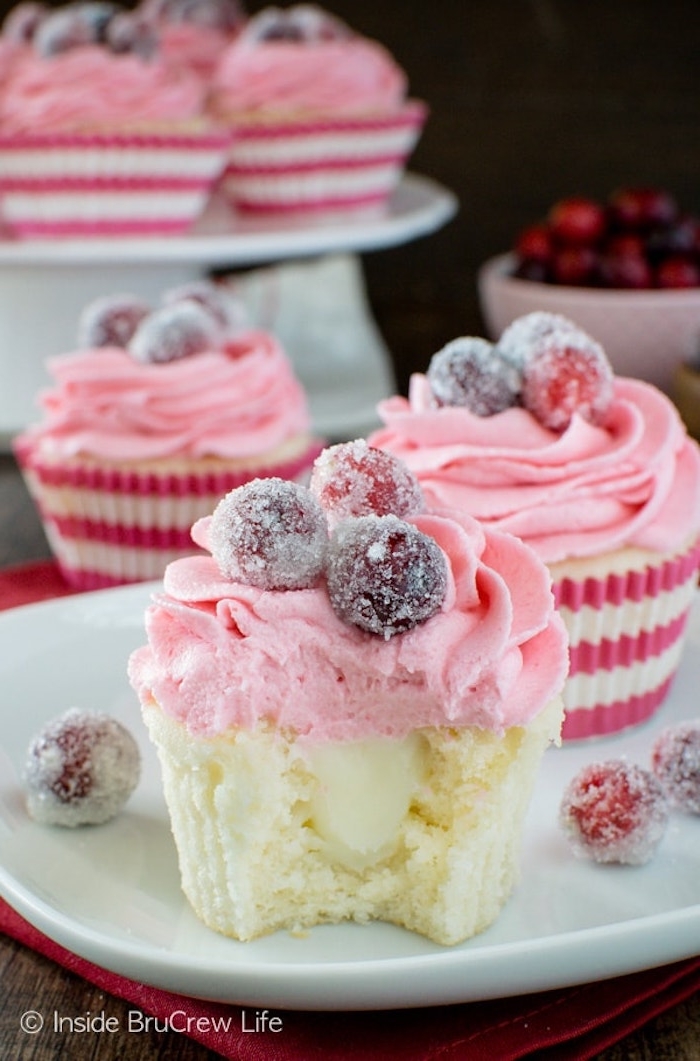 several pale yellow cupcakes, with pale pink icing, decorated with frosted cranberries, on white plate with more frosted cranberries