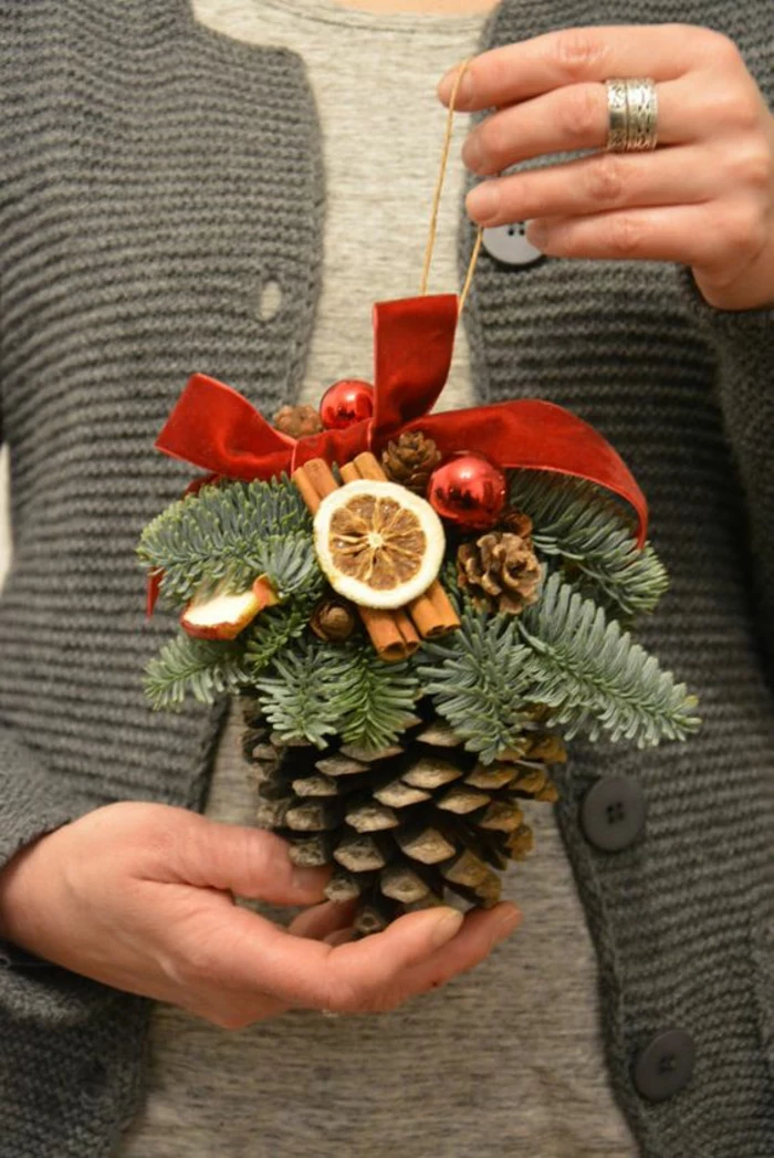 woman in grey sweater and light grey top, holding a christmas ornament made from a big pine cone, decorated with cinnamon sticks pine twigs and dried fruit, small ornaments and red ribbon