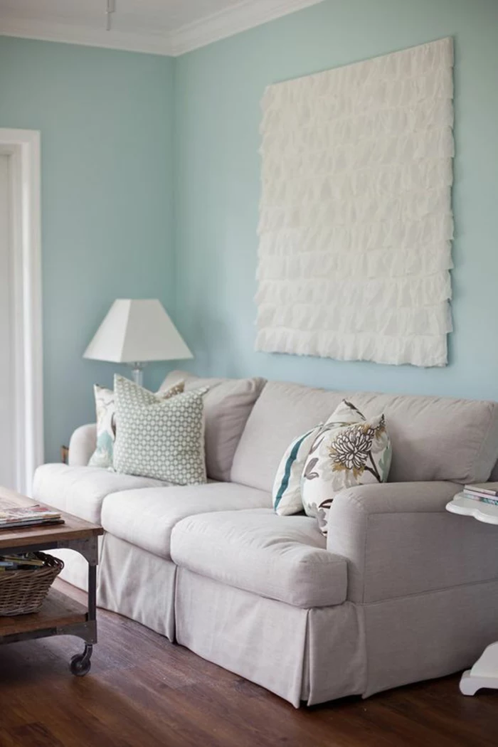 pale pastel blue wall, white ceiling plaster details and big square ruffled fabric ornament, pale grey couch with colorful cushions, white lamp and wooden table
