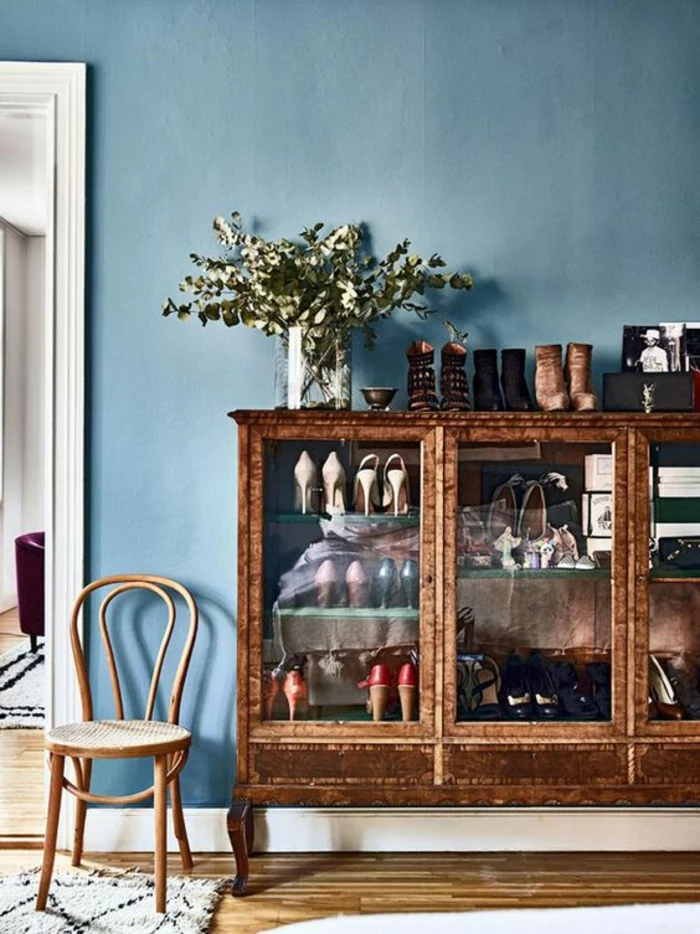 blue wall with white door frame, brown display cabinet containing many shoes, wooden chair and floors, glass vase with green plants, pale grey rug