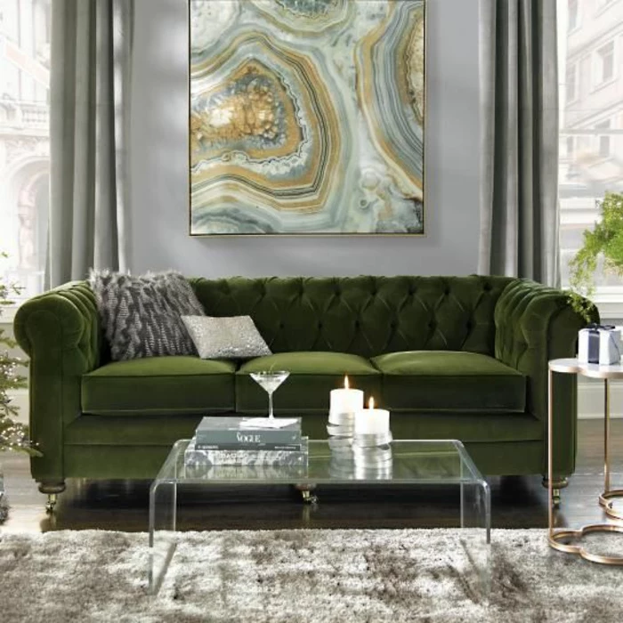 lush green sofa with two grey cushions, clear glass table with two lit candles, fluffy grey carpet, grey wall with yellow green and grey painting, windows with grey curtains
