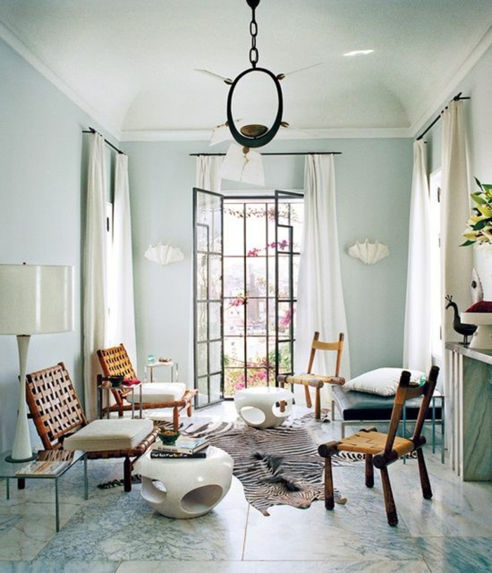 room with pale blue walls, windows and cream curtains, four wooden chairs and two small white tables, blue and cream tiled floor, white lamp and unusual chandelier, striped animal hide on floor