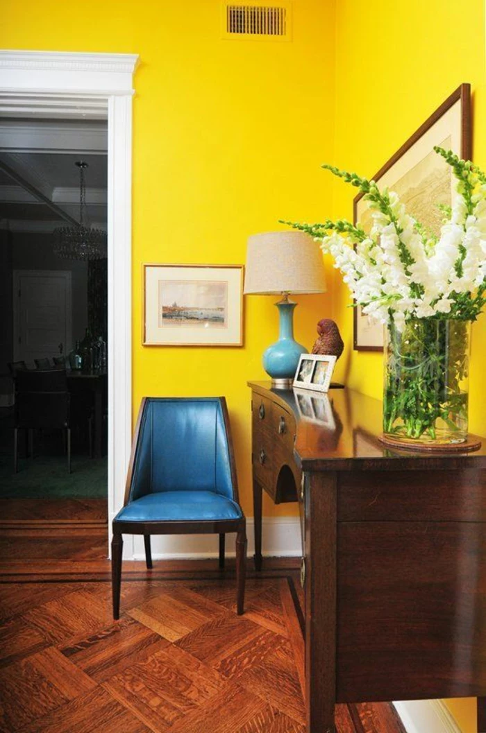 living room paint colors, bright yellow wall with white door frame, saturated blue chair and blue and cream lamp, brown wooden chest of drawers, wooden floor and big clear vase with white flowers