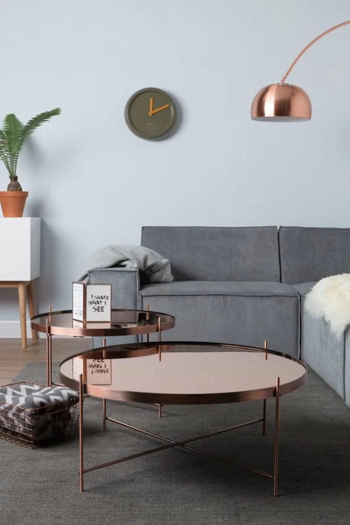 pale blue wall, grey sofa and white cupboard with wooden legs and potted plant, copper colored metal tables, grey carpet and wooden floor, olive green wall clock, grey blankets and cream sheep hide