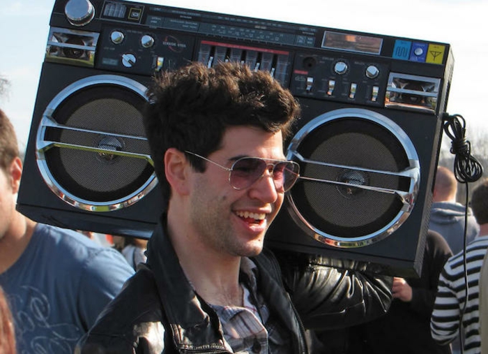 smiling man with gelled up wavy brown hair, aviator sunglasses and black leather jacket, carrying a retro stereo boombox, crowd in background