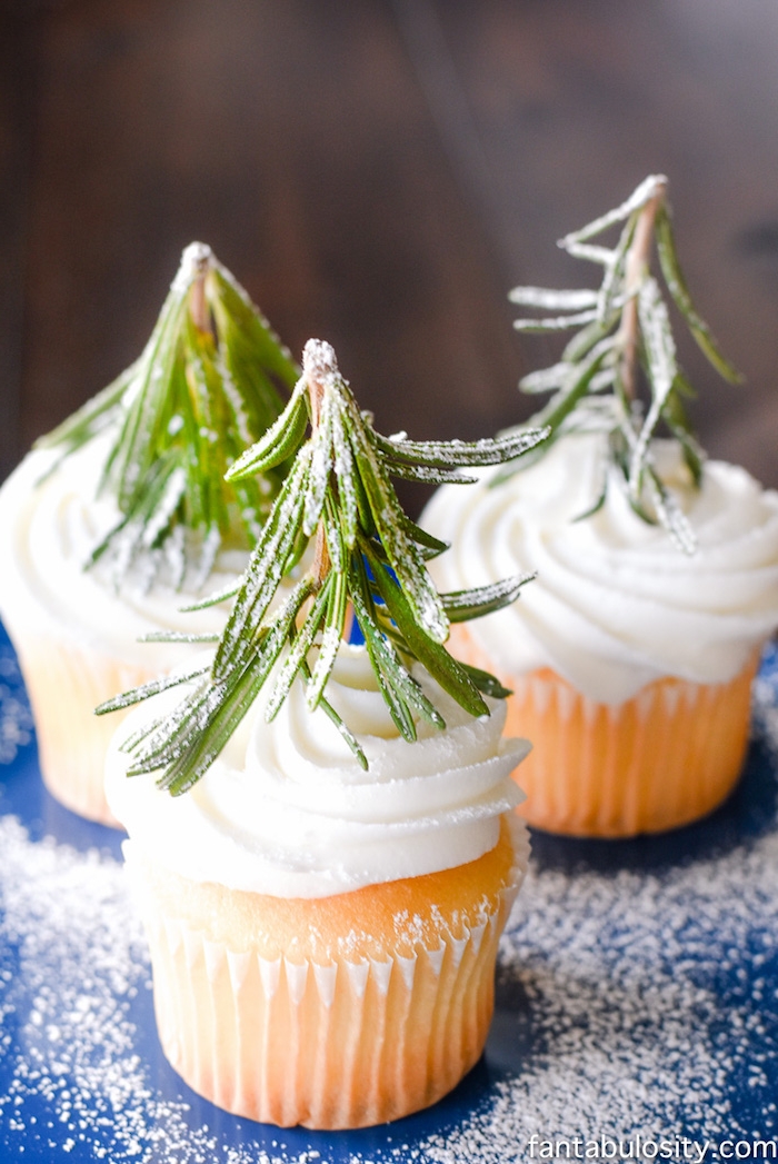 christmas flavors, three vanilla cupcakes with creamy white icing, each decorated with rosemary stalk and dusted with powdered sugar