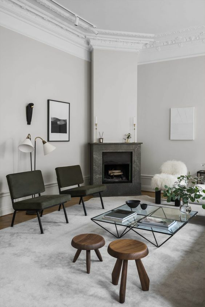 living room color schemes, pale grey walls and white ceiling with white plaster details, grey fireplace and two grey chairs, two small round wooden chairs, clear glass table with black metal legs and details