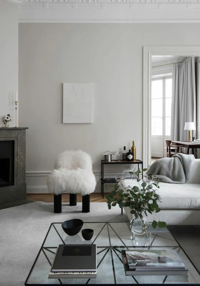 living room color schemes, white and pale grey room, dark grey fireplace, white fluffy chair with black legs, white sofa with grey blanket, clear glass table with black metal details