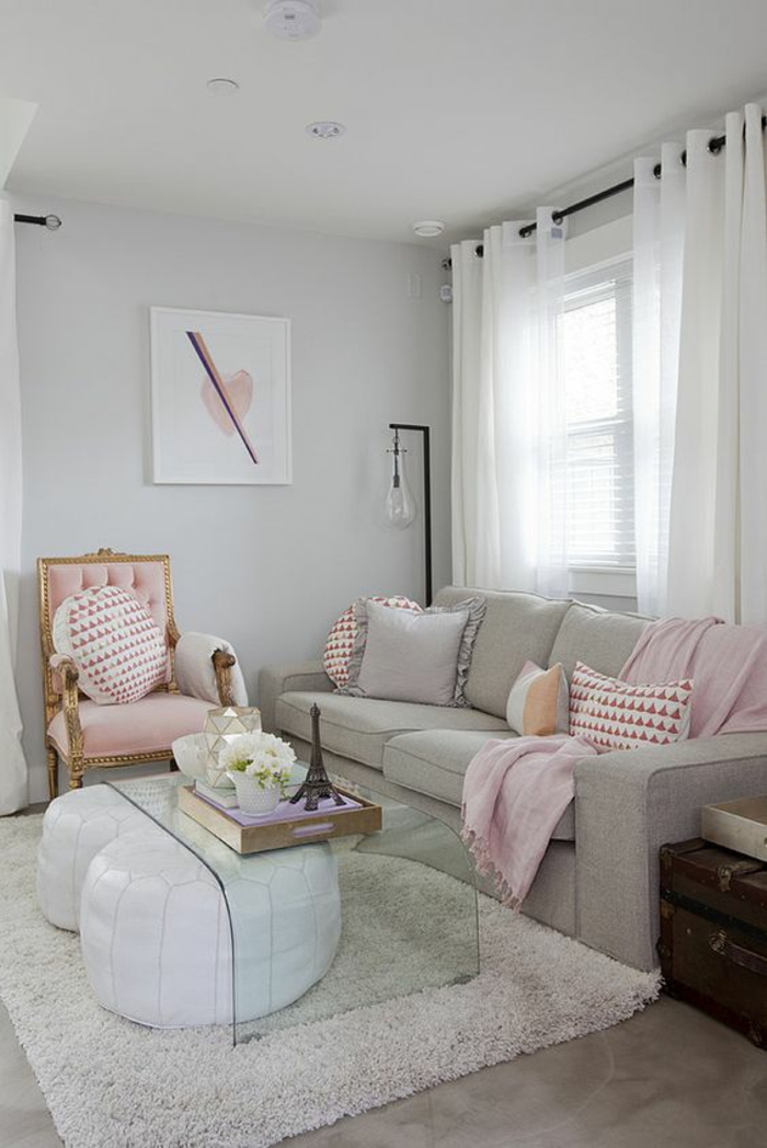 pale pink chair with gold details, light grey sofa with cushions and pink blanket, clear glass table and two white bean floor cushions, fluffy cream rug, window with white curtains
