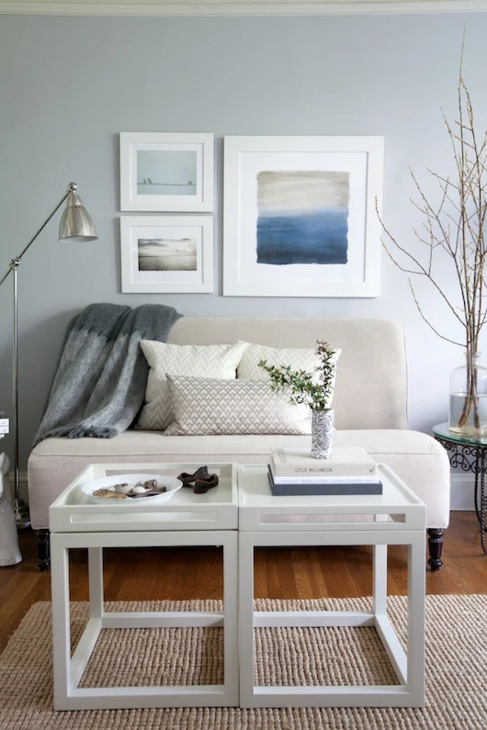 pale blue wall with three images in white frames, pale cream sofa with grey blanket and three pillows, small white table, wooden floor and cream rug